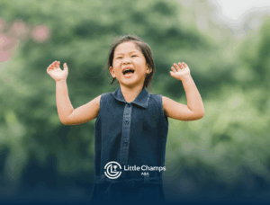 Smiling young child with arms raised, laughing happily outdoors against a soft green, natural background, blurred.