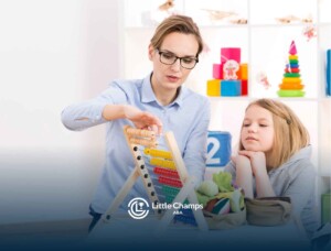 A teacher demonstrates an abacus to an attentive young girl in a bright classroom. Shelves with colorful toys and educational materials are in the background.