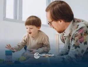 Young boy smiling while building with blocks on the floor beside a supportive ABA therapist.