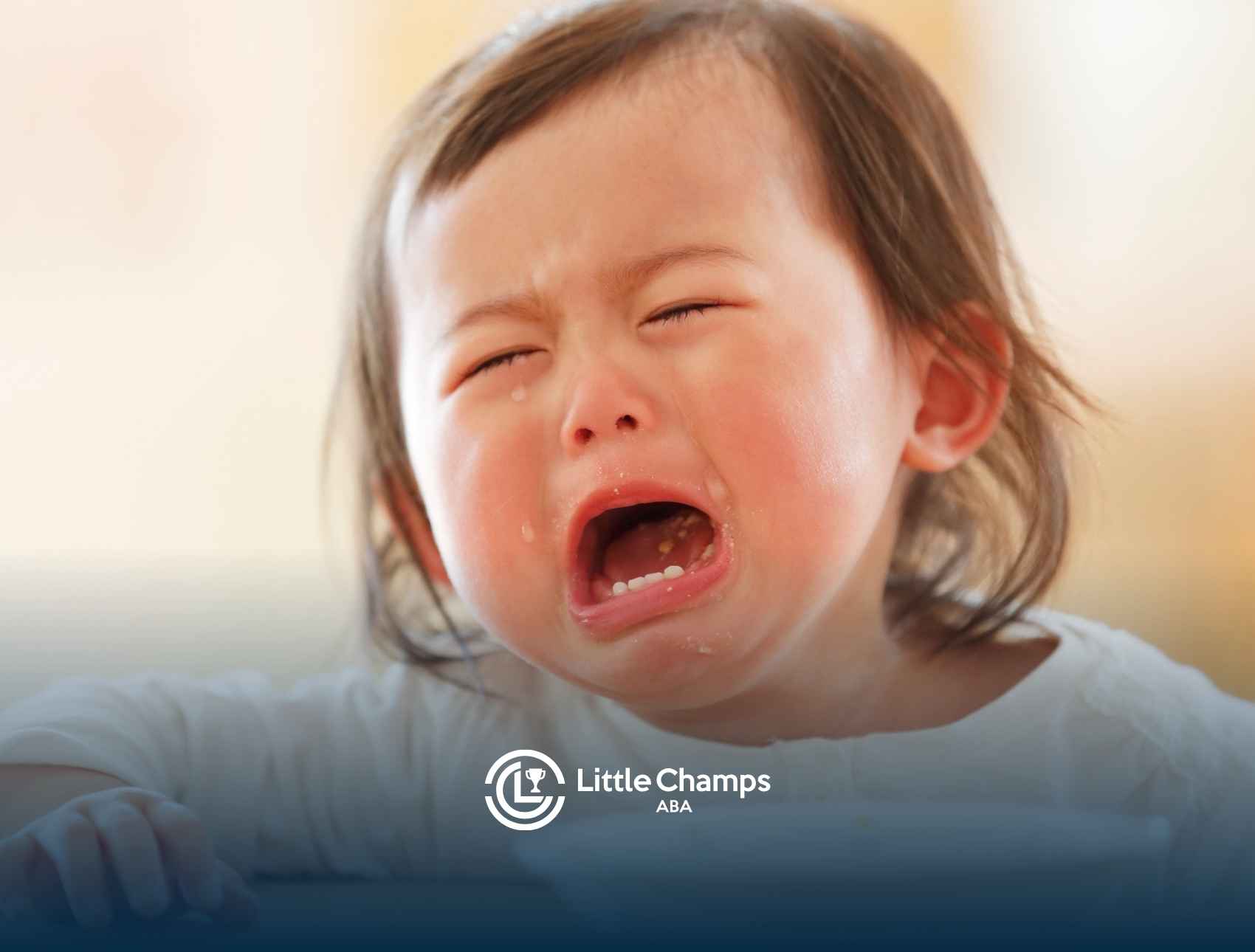 A toddler with brown hair is crying, eyes closed, and mouth open, wearing a white shirt. The image conveys distress.