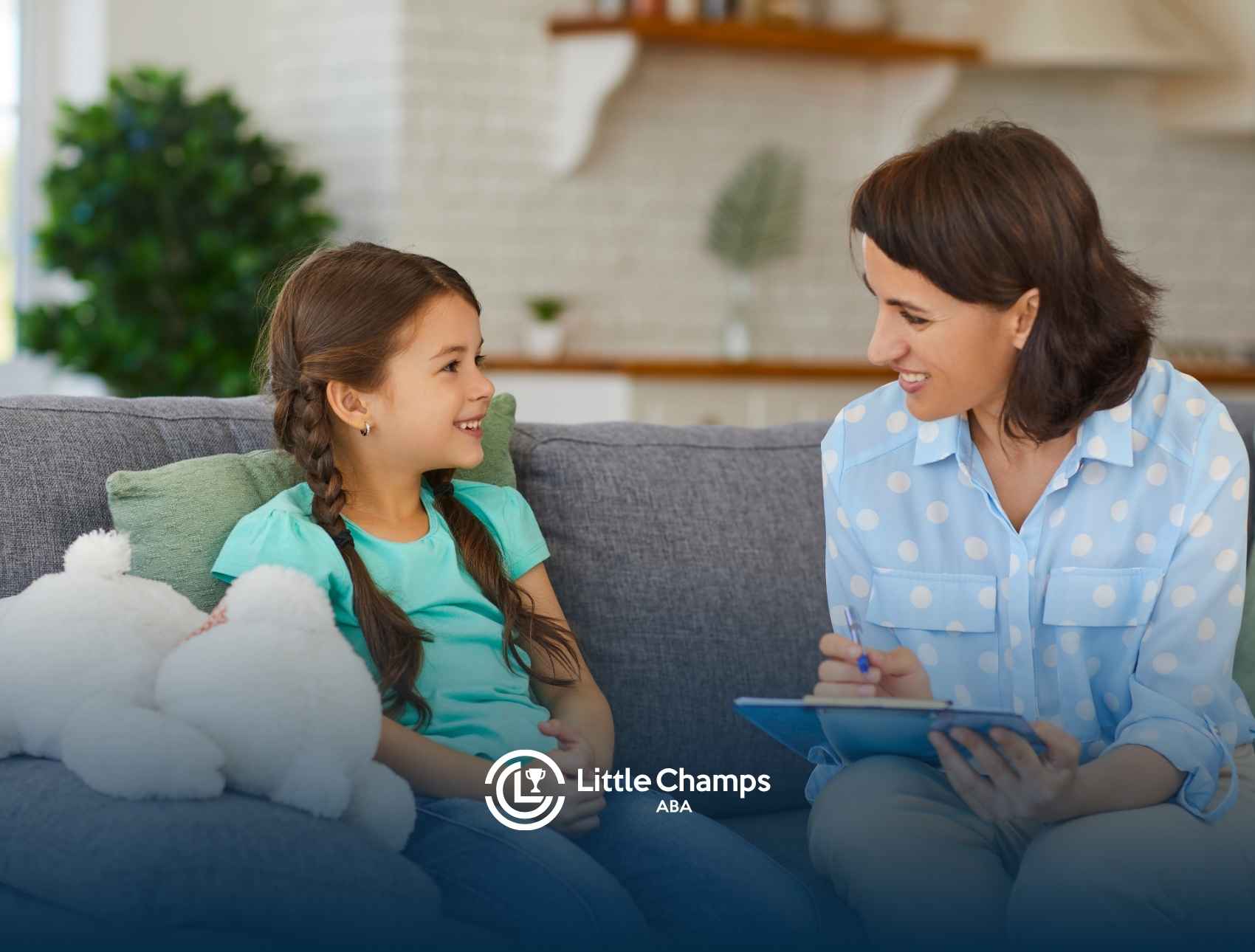 A woman in a blue polka dot shirt smiles and holds a clipboard while sitting on a couch with a young girl in a green shirt, conveying warmth and care.