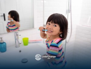 A young child with short dark hair smiles while brushing her teeth in a bright bathroom.