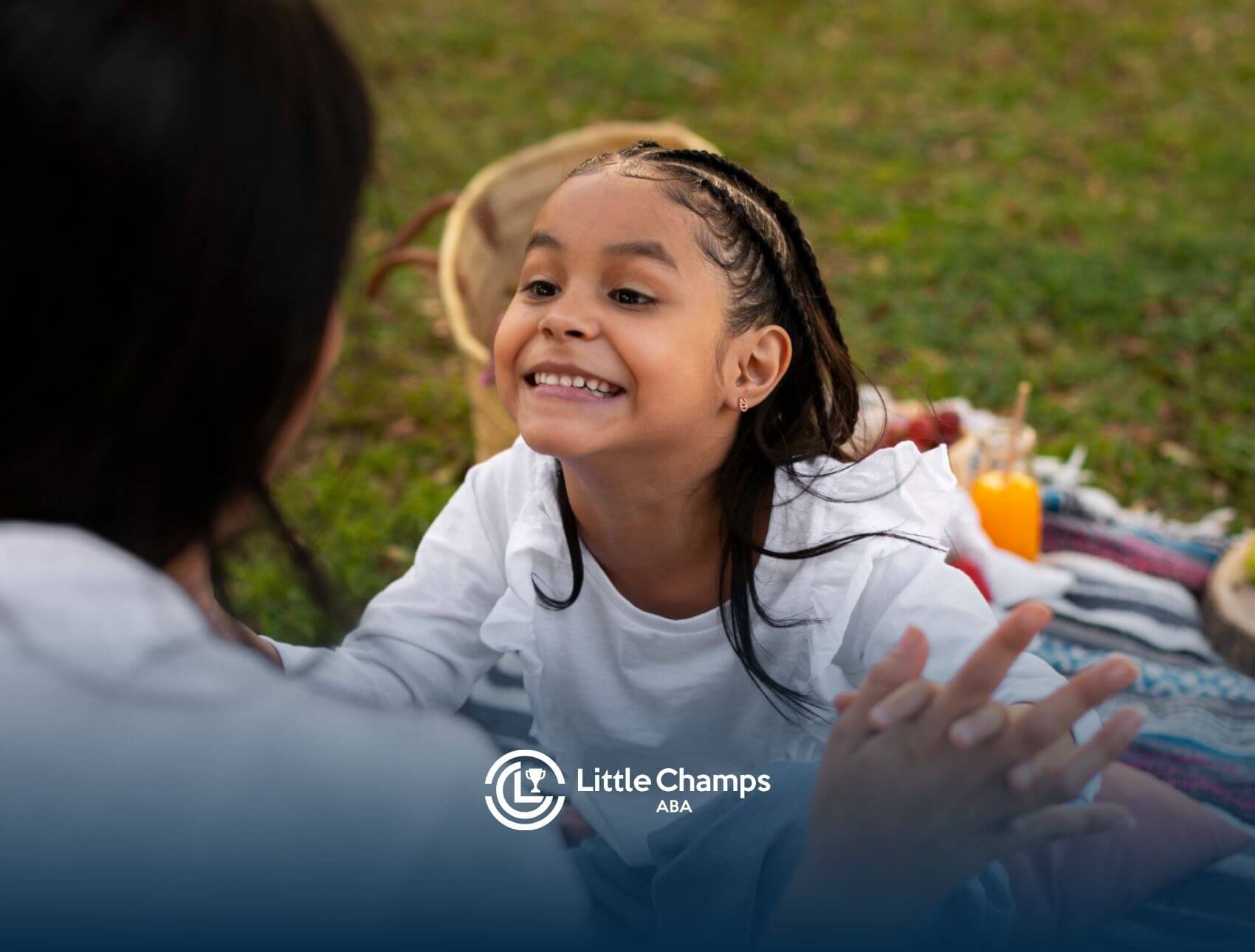 A young girl with autism smiles at ABA therapist while sitting on a picnic blanket outdoors.