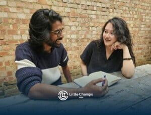 A man and woman sit at a wooden table outdoors, smiling and talking.