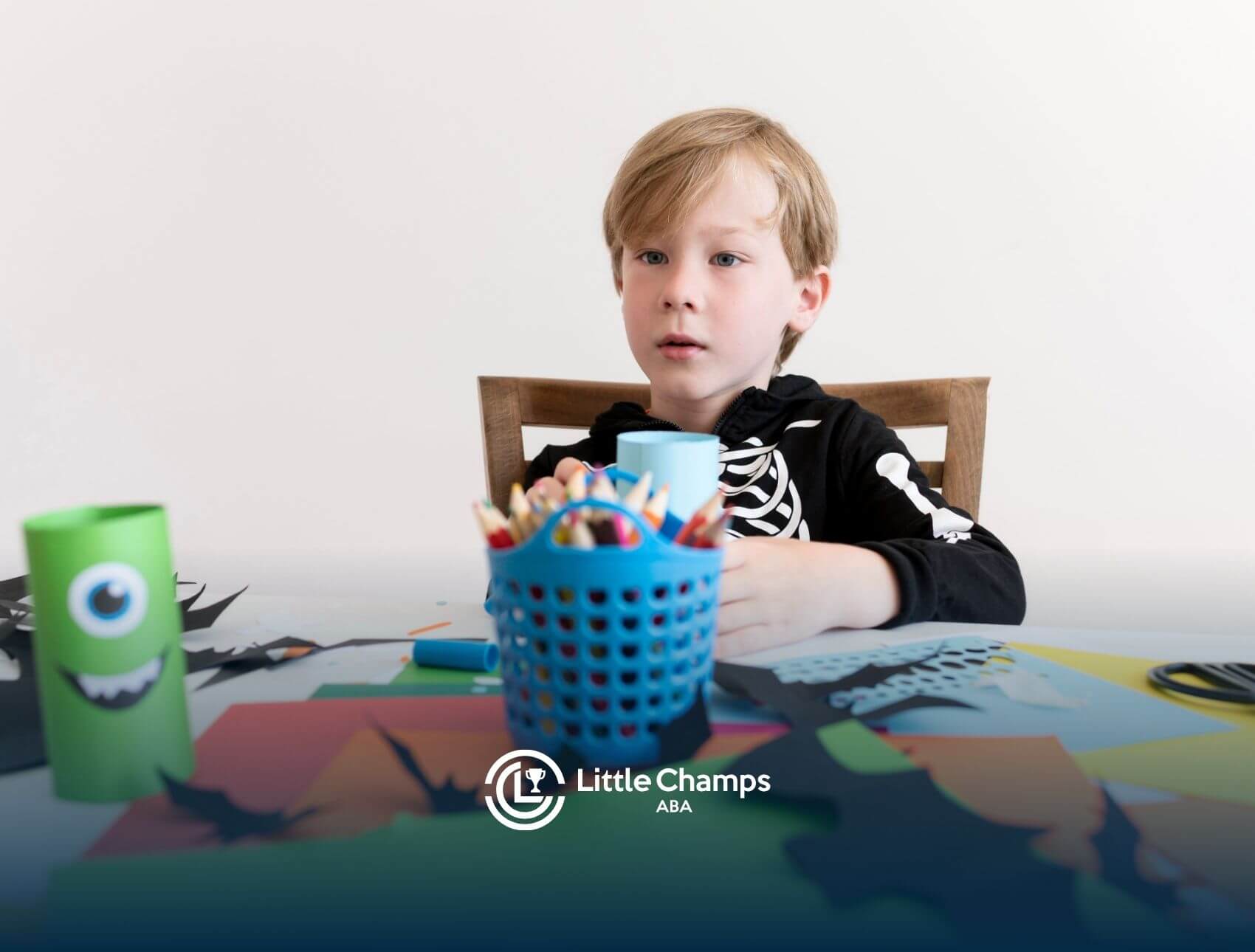 A young boy wearing a skeleton hoodie, sitting at a table surrounded by art supplies, looking focused.