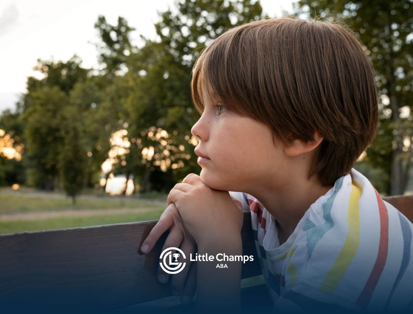 An autistic boy sitting on a park bench with his hands clasped, staring thoughtfully into the distance.