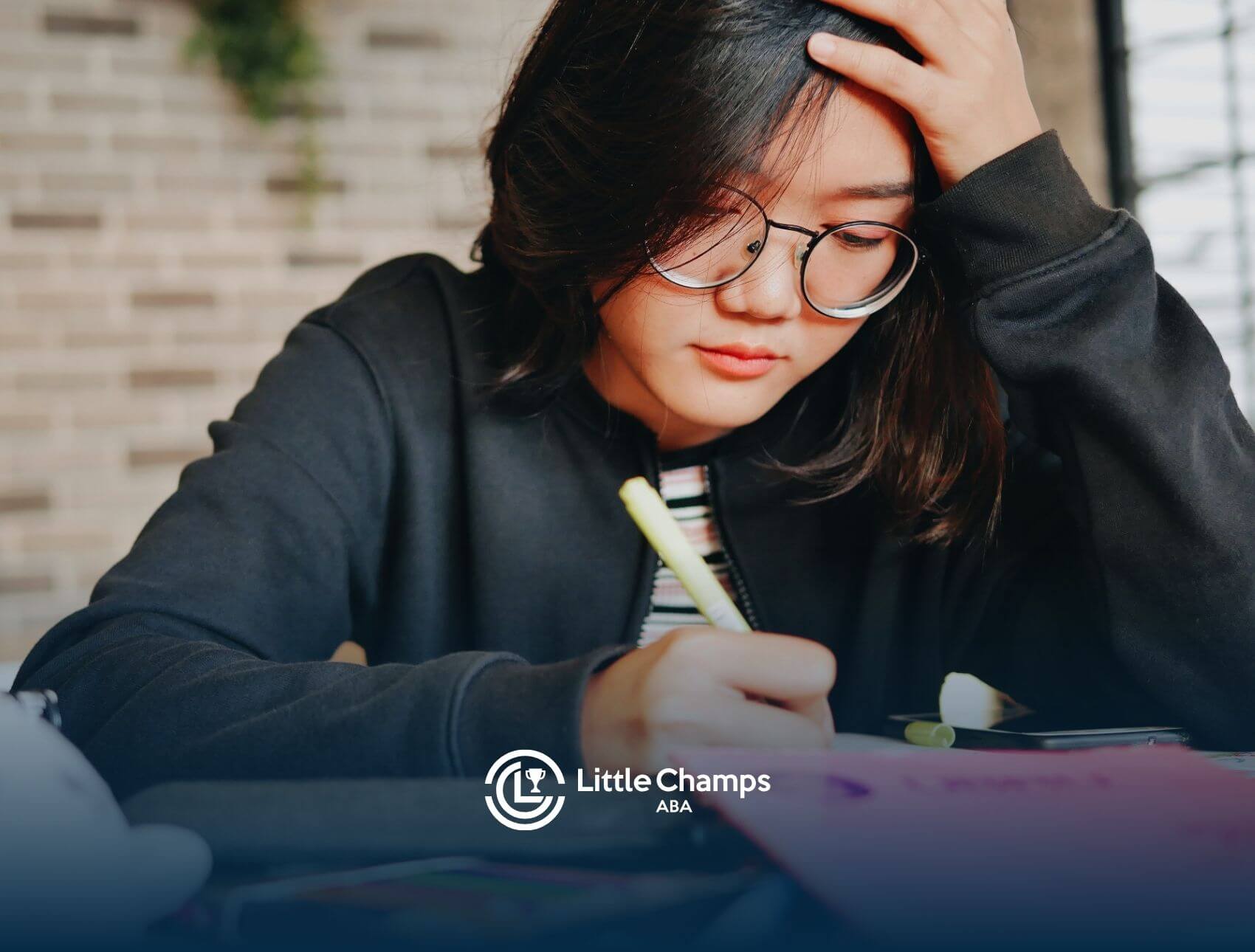 A teenage girl wearing glasses sits at a desk, pencil in hand, appearing stressed as she struggles with her work.
