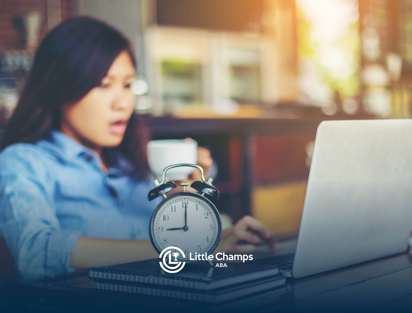 An ABA therapist working at her desk with a cup of coffee, an alarm clock, and a laptop, looking stressed.