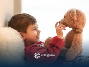 Smiling child in a red sweater lying by a window, playing with a teddy bear in ABA therapy.
