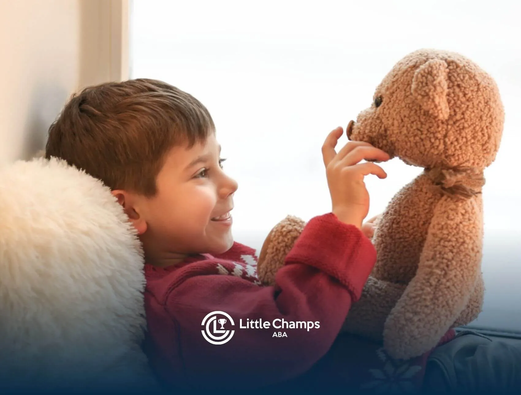 Smiling child in a red sweater lying by a window, playing with a teddy bear in ABA therapy.