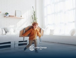 Toddler wearing cardboard wings running indoors in a bright living room.