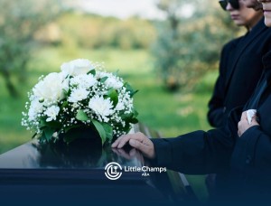 A person in black funeral attire resting a hand on a wooden casket topped with white flowers.