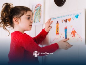 A young girl with autism hanging a colorful drawing of rockets and hearts in a white frame on a wall.