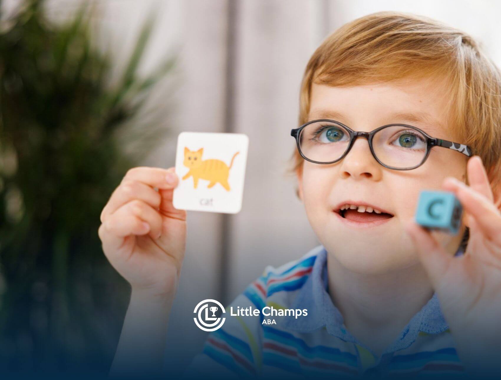 An autistic boy with glasses smiling while holding up a "cat" flashcard.